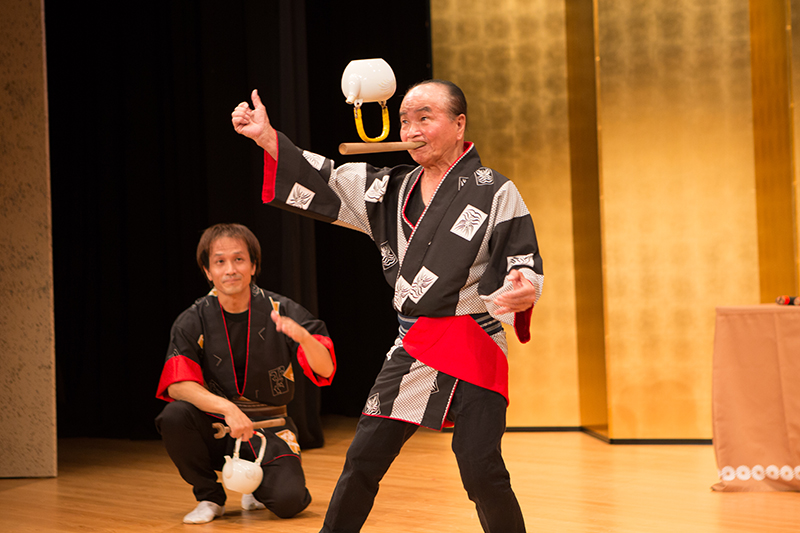 ステージで土瓶の曲の芸を披露するラッキー師匠と傍らで見つめる豊来家幸輝